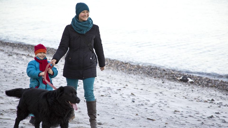 Eine Frau geht mit einem Kind und einem schwarzen Hund am Strand spazieren