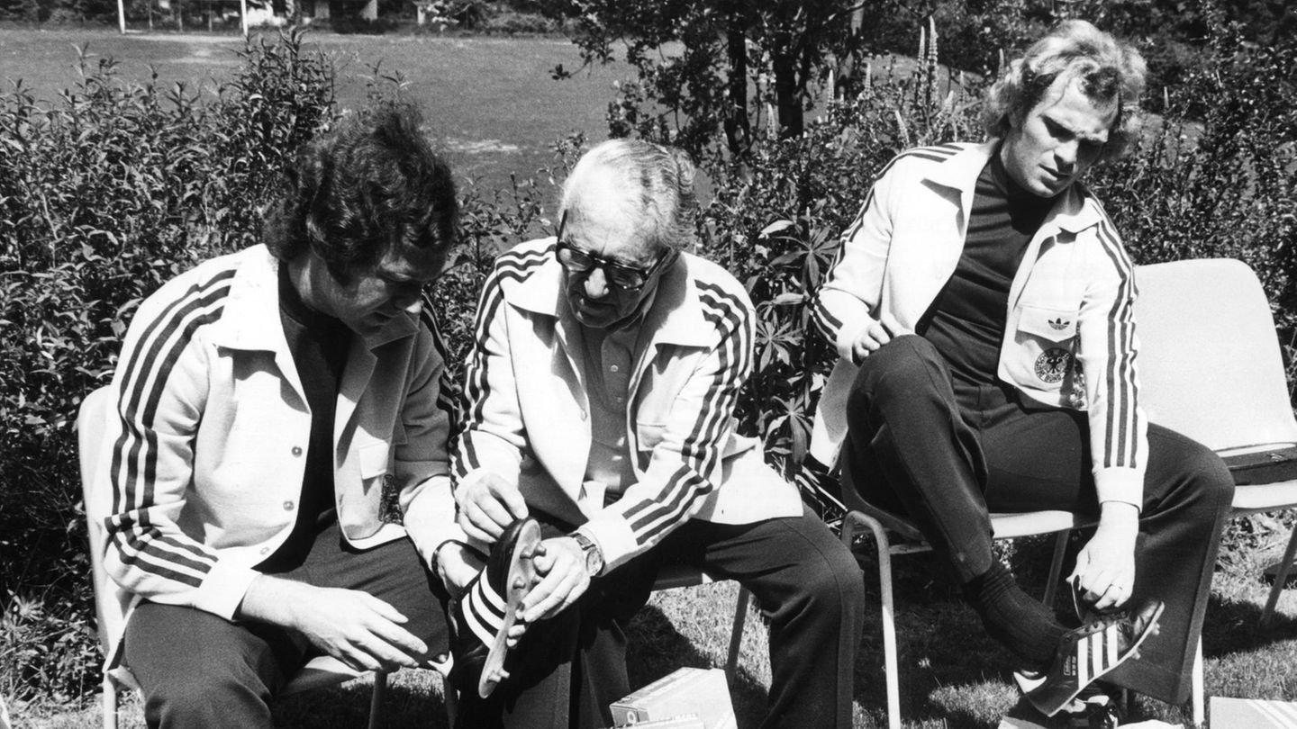 Vor der WM 1974 probieren Libero und Kapitän Franz Beckenbauer (l.) und Mittelfeldspieler Uli Hoeneß (r.) Fußballschuhe an, die Adidas-Gründer Addi Dassler (M.) persönlich in der Sportschule Malente (Schleswig-Holstein) abliefert.