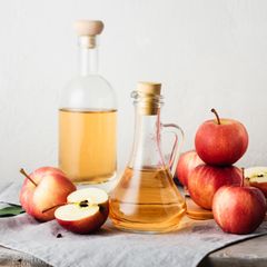 Apfelessig: Zwei Flaschen mit Apfelessig stehen auf einem Tisch, daneben liegen Äpfel