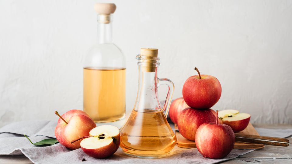 Apfelessig: Zwei Flaschen mit Apfelessig stehen auf einem Tisch, daneben liegen Äpfel