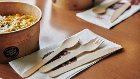 Vorne Holzbesteck auf Serviette und links daneben ein Behälter mit einer Suppe auf einem Holztisch