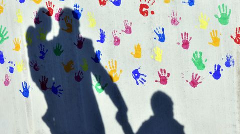 Ein Mann mit einem Kind auf dem Arm und einem an der Hand wirft einen Schatten auf eine mit bunten Handabdrücken bemalte Wand