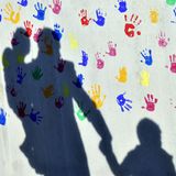 Ein Mann mit einem Kind auf dem Arm und einem an der Hand wirft einen Schatten auf eine mit bunten Handabdrücken bemalte Wand