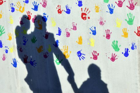 Ein Mann mit einem Kind auf dem Arm und einem an der Hand wirft einen Schatten auf eine mit bunten Handabdrücken bemalte Wand