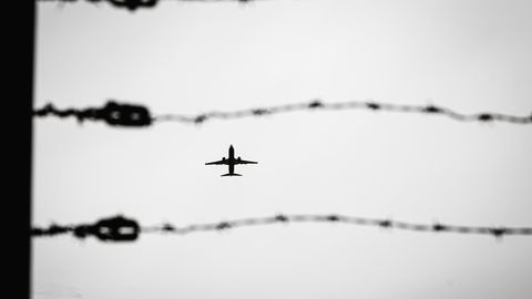 Durch eine Stacheldrahtzaun sieht man ein Passagierflugzeug am Himmel