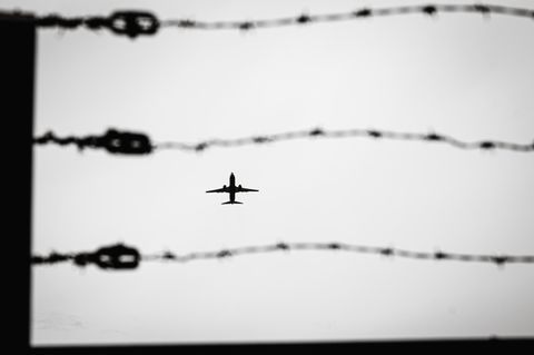 Durch eine Stacheldrahtzaun sieht man ein Passagierflugzeug am Himmel