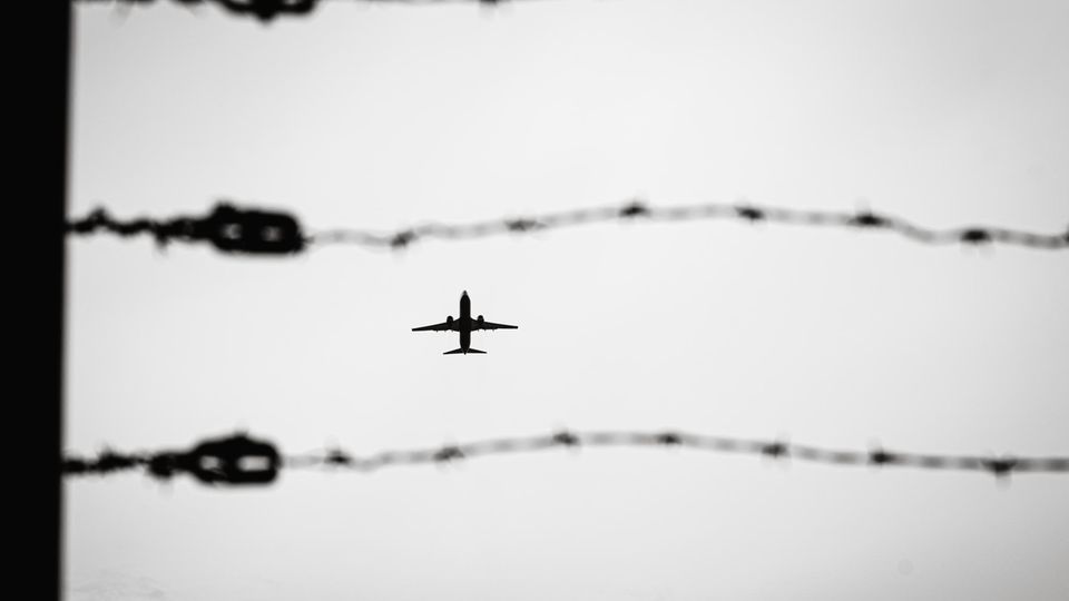 Durch eine Stacheldrahtzaun sieht man ein Passagierflugzeug am Himmel