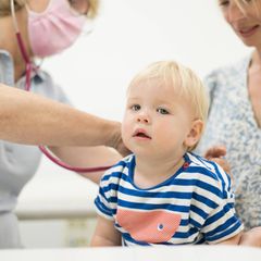 Blondes Kleinkind sitzt bei erwachsener Person auf dem Schoß und wird von Ärztin mit Stethoskop untersucht
