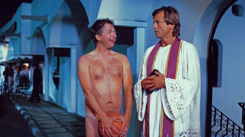 Ein nackter Mann steht neben einem Priester und hält sich einen Ball vor sein Geschlechtsorgan.