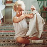 Ein fünfjähriges Mädchen im Unterhemd sitzt im Schneidersitz auf dem Boden und zerschneidet einen Pullover.