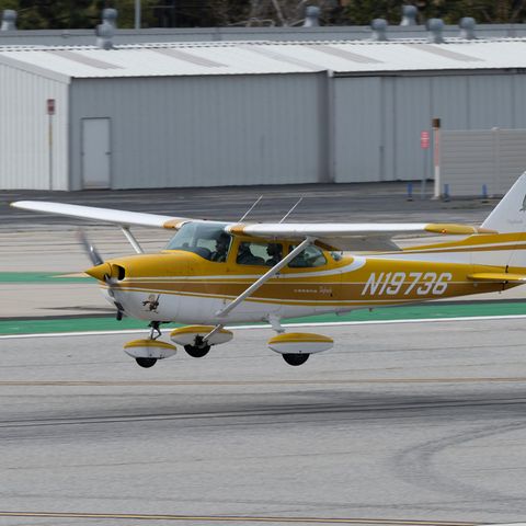 Eine goldgelb und weiß lackierte Cessna 172 fliegt tief über einer Start- und Landebahn