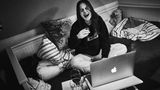 Janne sitzt lachend mit einem Glas Wein vor ihrem Laptop für ein virtuelles Treffen mit ihren Freunden.