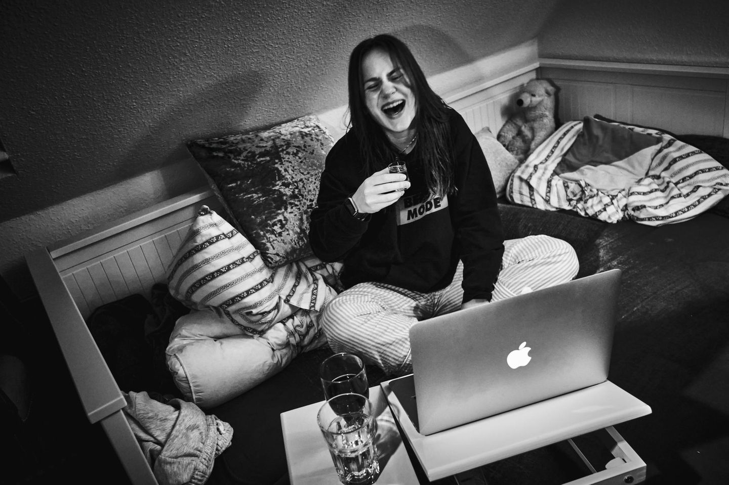 Janne sitzt lachend mit einem Glas Wein vor ihrem Laptop für ein virtuelles Treffen mit ihren Freunden.