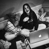 Janne sitzt lachend mit einem Glas Wein vor ihrem Laptop für ein virtuelles Treffen mit ihren Freunden.