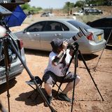 Mann guckt sitzend auf Parkplatz durch Kamera mit Sonnenschutz