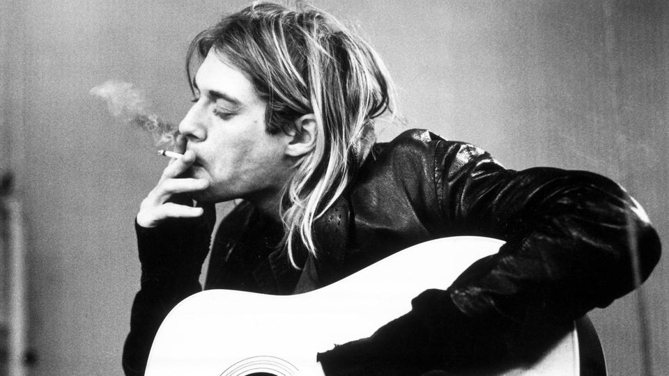 Kurt Cobain zieht mit geschlossenen Augen an einer Zigarette, während sein linker Arm über seiner Gitarre ruht