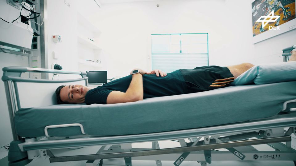 Ein Mensch liegt für die Weltraum-Studie auf einem Krankenhausbett, leicht geneigt und lächelt in die Kamera, er trägt Shorts