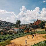 Kinder spielen auf einem Sandplatz. Im Hintergrund Häuser und Hügel