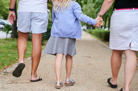 Symbolbild Namensrecht: Ein Vater und eine Mutter halten die Hände ihrer Tochter, während sie auf einem Gehweg gehen