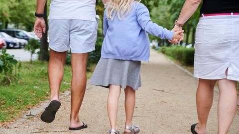 Symbolbild Namensrecht: Ein Vater und eine Mutter halten die Hände ihrer Tochter, während sie auf einem Gehweg gehen