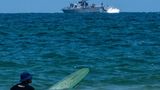 Ein Surfer wartet auf eine Welle, während ein israelisches Marineschiff im Mittelmeer vor der Küste patrouilliert