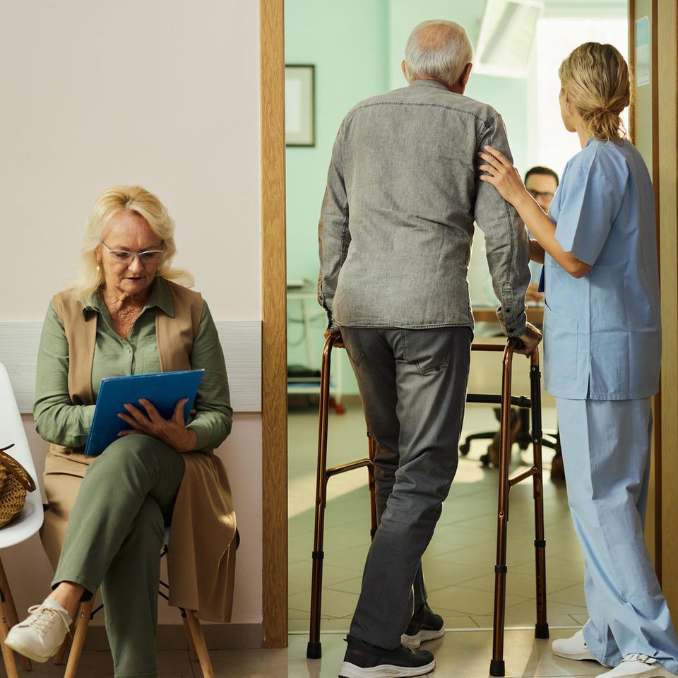 Im Inneren einer Hausarztpraxis: Menschen warten auf Stühlen, während ein Patient ins Behandlungszimmer geführt wird.