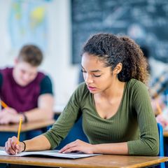 Junge Frau sitzt mit Mitschülern im Klassenraum und schreibt auf ein Aufgabenblatt