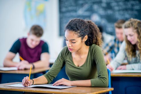 Junge Frau sitzt mit Mitschülern im Klassenraum und schreibt auf ein Aufgabenblatt