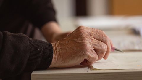 Die rechte Hand eines alten Mannes hält einen Kugelschreiber über ein weißes Blatt Papier