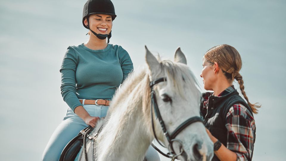 Eine lachende Frau sitzt auf einem Pferd, eine weiter hält die Zügel fest und guckt ernst