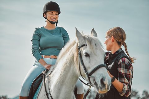 Eine lachende Frau sitzt auf einem Pferd, eine weiter hält die Zügel fest und guckt ernst
