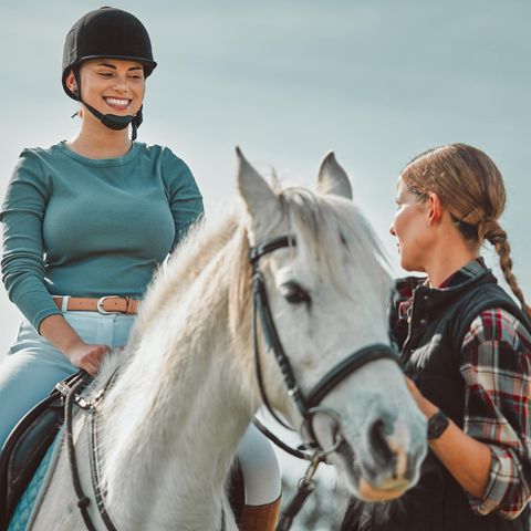 Eine lachende Frau sitzt auf einem Pferd, eine weiter hält die Zügel fest und guckt ernst