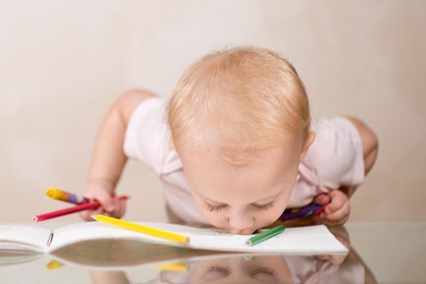 Ein Kleinkind mit Buntstiften innder Hand beißt in ein Buch