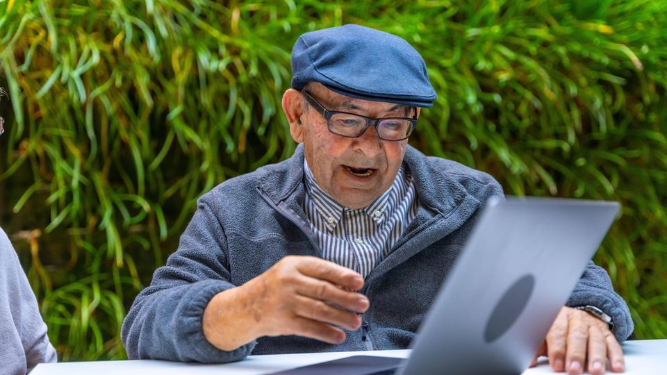 Ein älterer Herr mit Mütze und Brille sitzt lachend vor einem Laptop