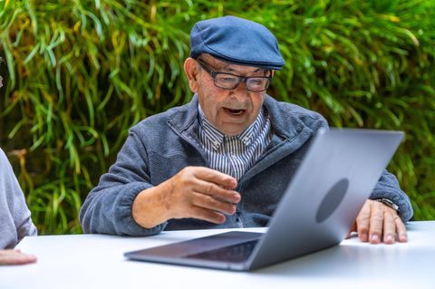 Ein älterer Herr mit Mütze und Brille sitzt lachend vor einem Laptop