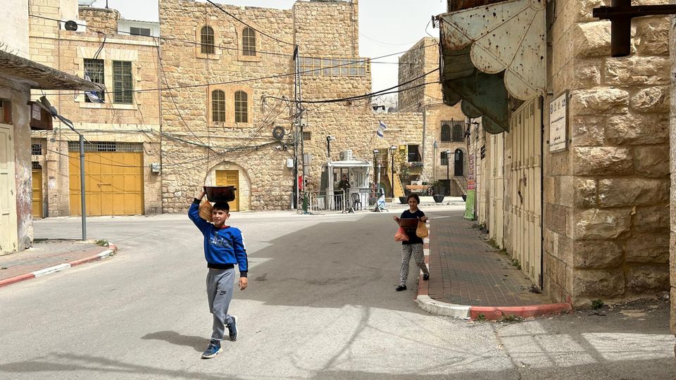 Die Geschwister Malik (vorne) und Melek (hinten) auf dem Heimweg von der Suppenküche. Im Hintergrund ein israelischer Checkpoint, bewacht von einem Soldaten, der auch Siedler ist.