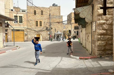 Die Geschwister Malik (vorne) und Melek (hinten) auf dem Heimweg von der Suppenküche. Im Hintergrund ein israelischer Checkpoint, bewacht von einem Soldaten, der auch Siedler ist.
