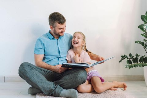 Ein Vater sitzt auf dem Boden und neben ihm seine Tochter, der er ein Buch vorliest.