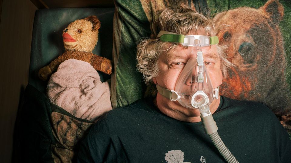 Torsten Herrmann (Doc Mabuse) in seinem Bett in der Reha-Einrichtung mit Atemmaske, daneben ein Teddybär