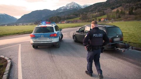 ein Polizist der Grenzpolizei hält vor einem Bergpanorama ein schwarzes Auto zur Kontrolle an