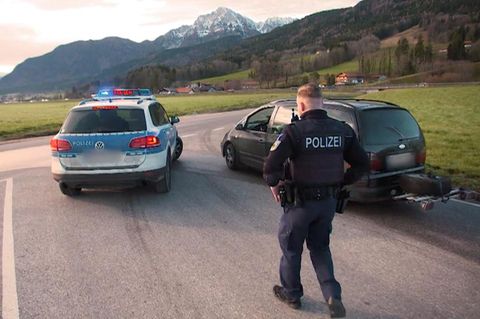 ein Polizist der Grenzpolizei hält vor einem Bergpanorama ein schwarzes Auto zur Kontrolle an