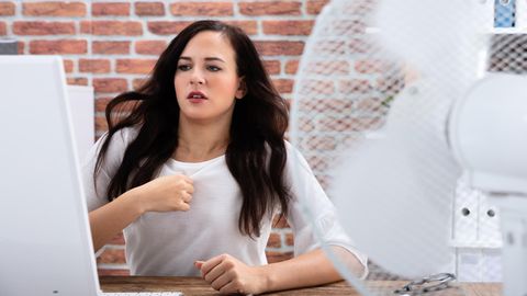 Eine Frau sitzt am Schreibtisch und veruscht sich mit einem Ventilator abzukühlen