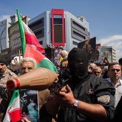 Soldat der Revolutionsgarde im Iran mit Sturmmaske steht in Menschenmenge und zielt mit seiner Panzerfaust auf die Kamera