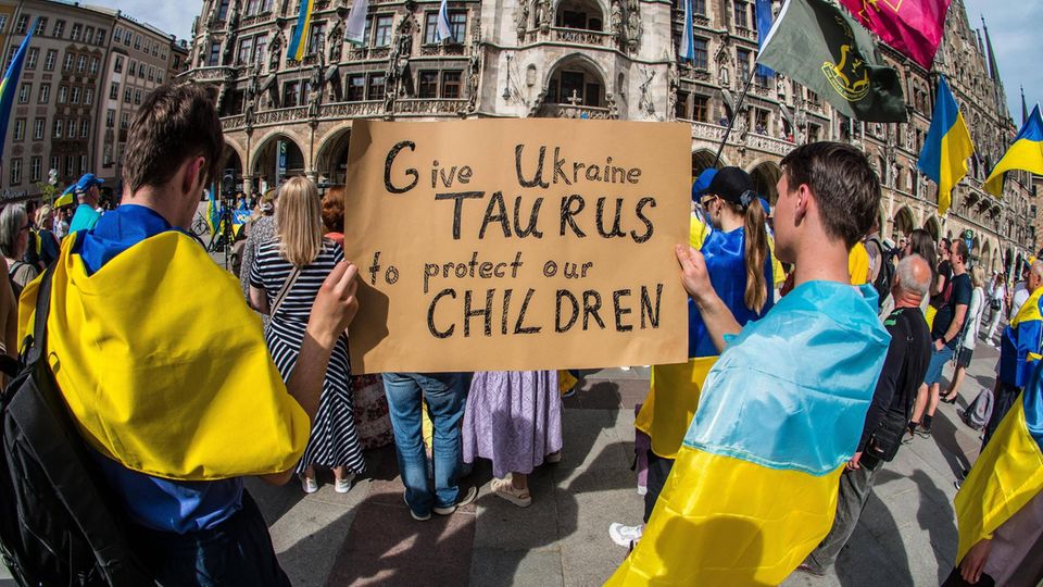 In München haben sich Menschen ukrainische Flaggen um den Hals gebunden und halten ein Schild hoch, auf dem sie die Lieferung fordern.