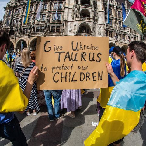 Menschen in München haben Ukraine-Fahnen umgebunden und halten ein Plakat, das die Lieferung fordert.