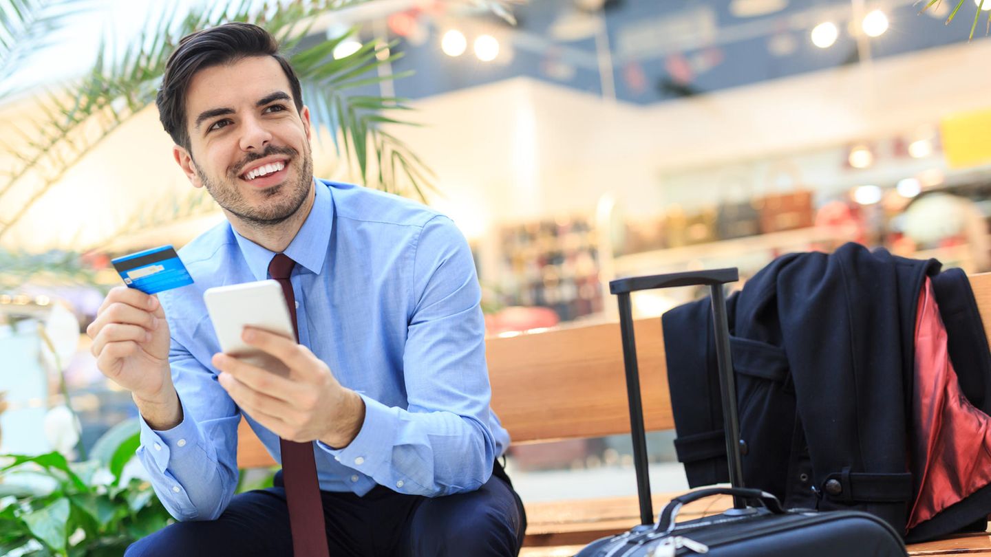 Premium-Kreditkarten: Wo winken die meisten Vorteile? Mann sitzt am Flughafen und hält lächelnd eine Kreditkarte in der Hand.