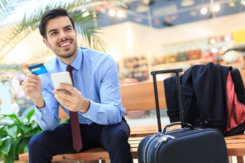 Mann sitzt am Flughafen und hält lächelnd eine Kreditkarte in der Hand.