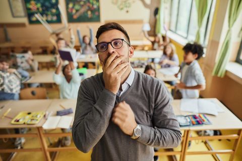 Lehrer mit Hand vor dem Mund vor unruhiger Schulklasse, in Deutschland herrscht Lehrermangel