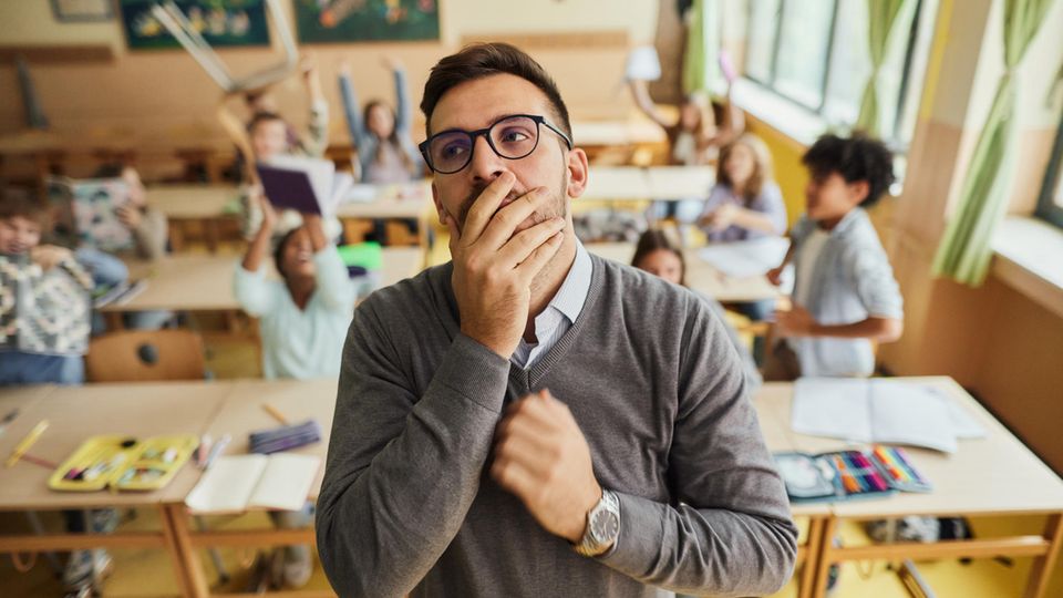 Lehrer mit Hand vor dem Mund vor unruhiger Schulklasse, in Deutschland herrscht Lehrermangel