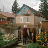 Eine ältere Dame steht an einem Gartentörchen und schaut auf die Straße.